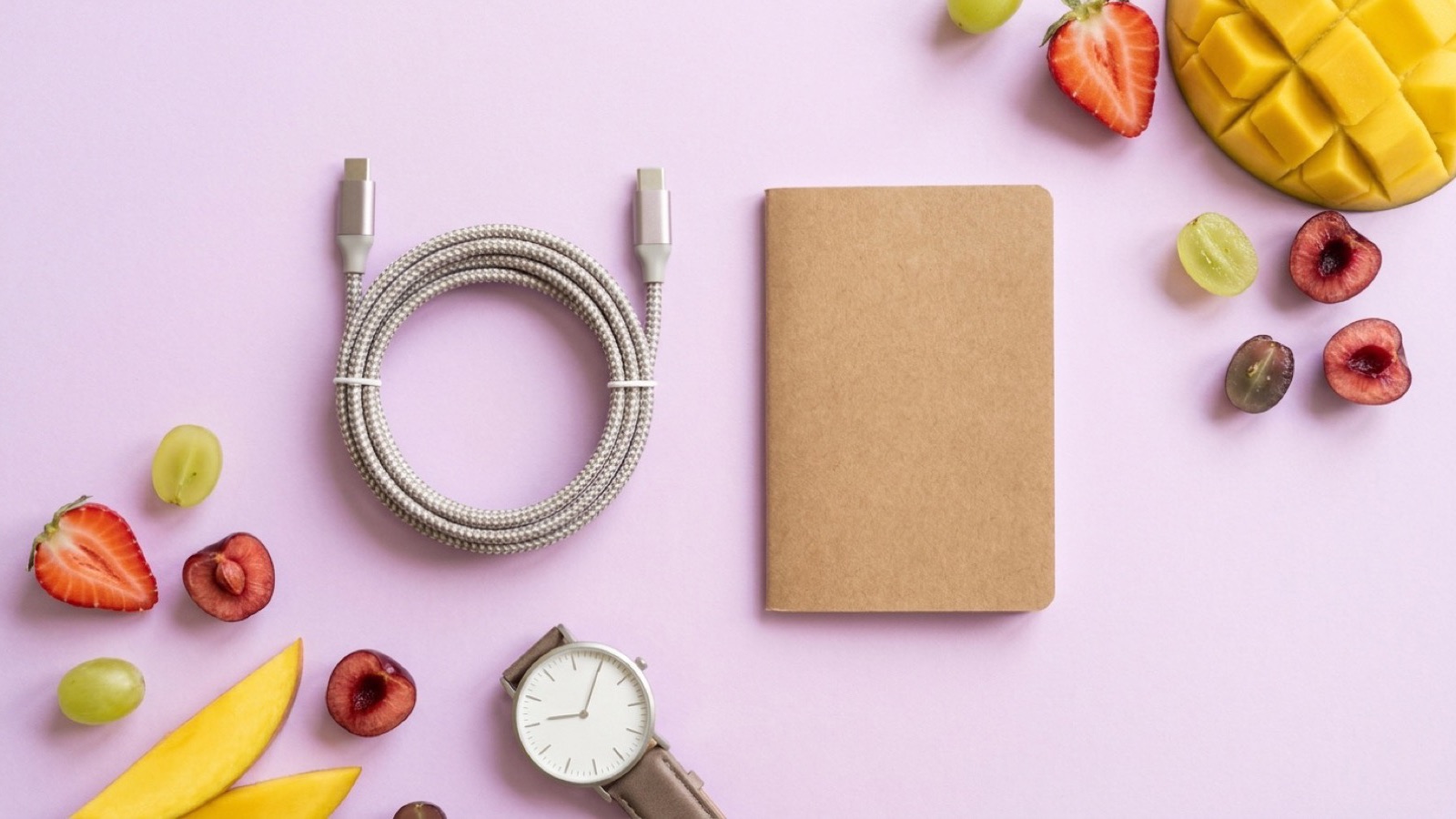 Flat lay s USB-C kabelem, zápisníkem, hodinkami a ovocem na fialovém pozadí